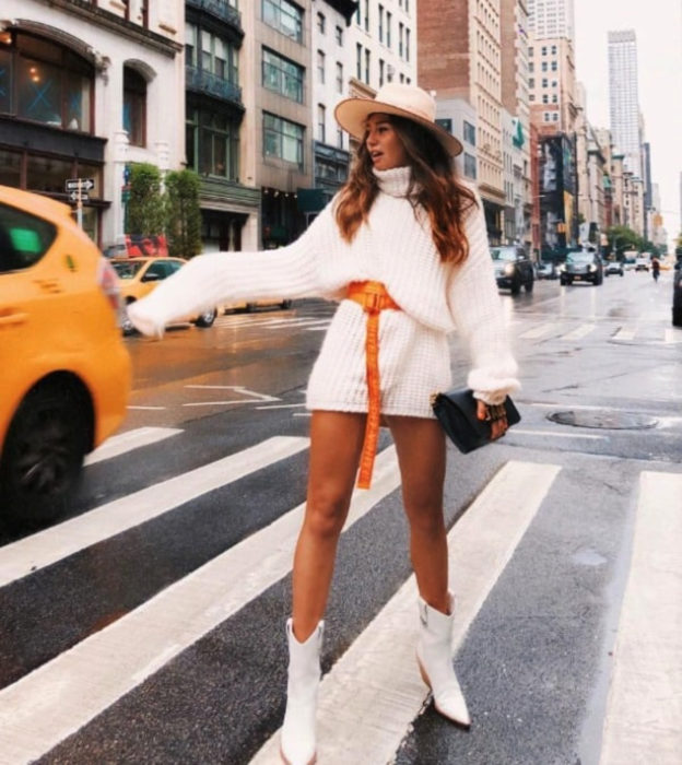 Chica en cruce peatonal haciéndole la parada a un taxi, usa suéter blanco grande como vestido corto de otoño, con cinturón anaranjado y botas puntiagudas y blancas, con sombrero y bolsa de mano