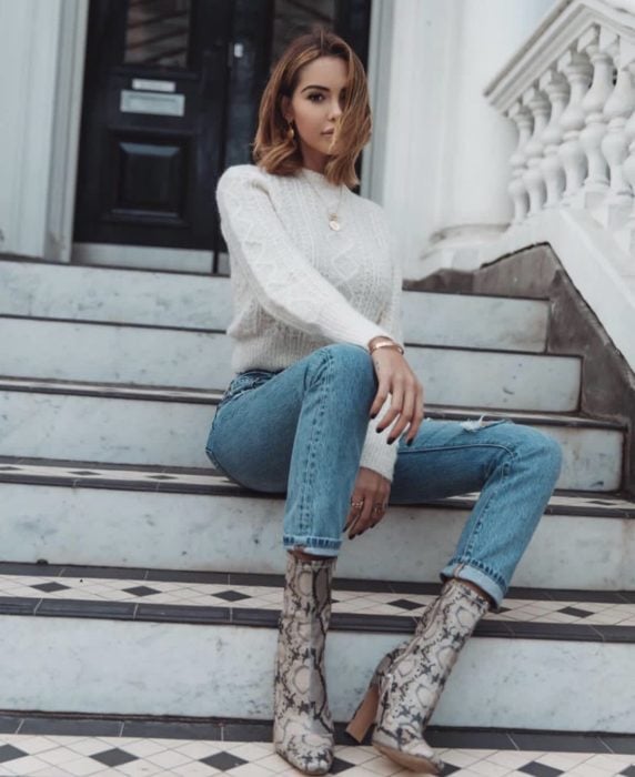 Chica sentada en unas escaleras durante una sesión de fotos mientras usa un sueter blanco, jeans y botas de estampado animal 