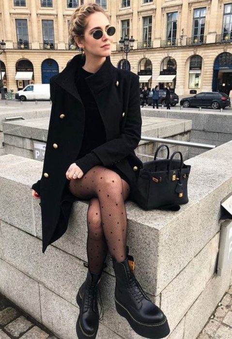 Chica sentada en una fuente mientras posa para una fotografía y usa un look negro con botines militares 