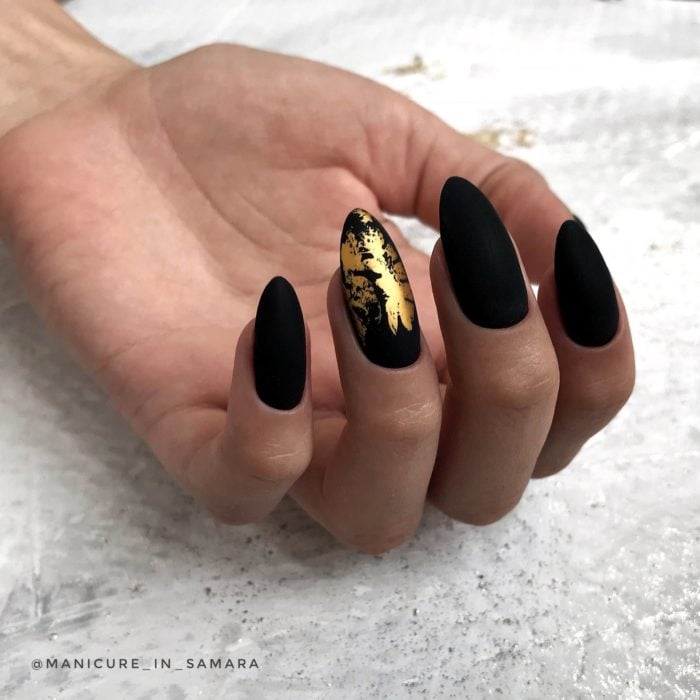 Uñas largas en forma de almendra color negro mate con una uña con detalles dorados