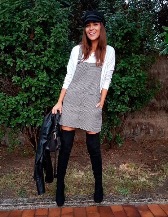 Chica usando un vestido pichi de color gris con botas y boina de color negro 