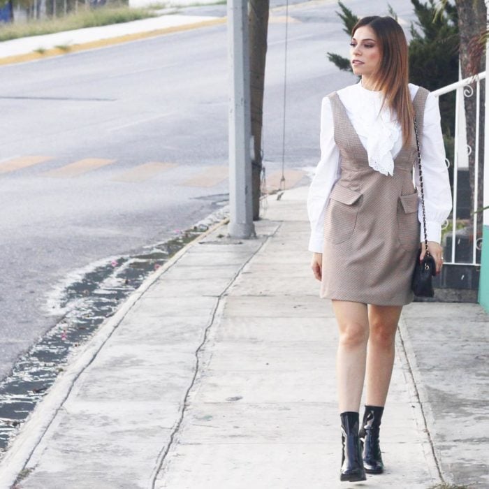 Chica caminando por la calle mientras está usando un vestido de color café y una blusa blanca combinados con unos botines negros 
