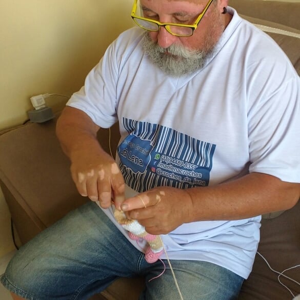 Joao Stanganelli tejiendo una muñeca, sentado en un sofá y usando lentes y una camiseta