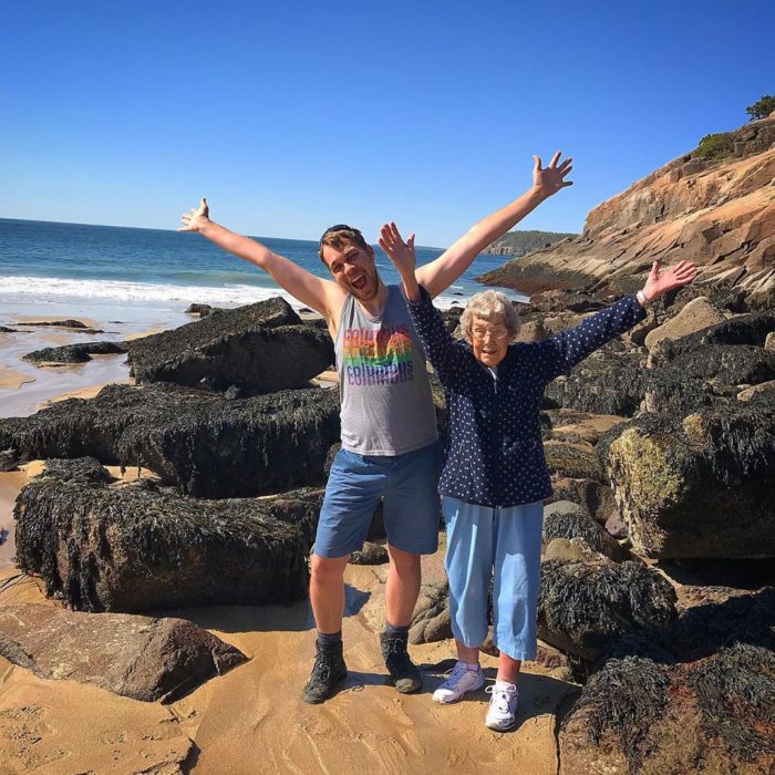 Joy y su nieto Brad en una playa con rocas con los brazos abiertos hacia arriba