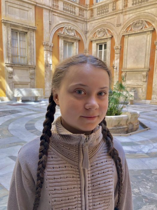 Greta Thunberg en un patio interior de un edificio con chamarra café y sus trenzas