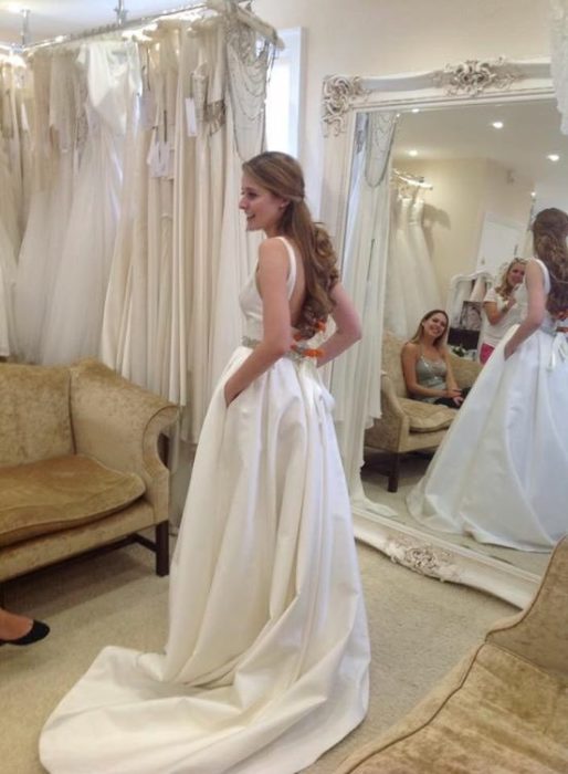 una mujer probándose un vestido de novia frente a un espejo en el que se reflejan otras personas que la observan