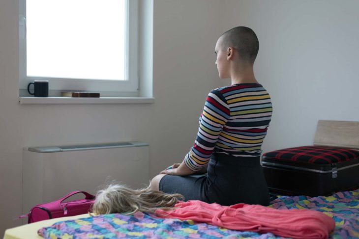 Mujer sentada en la orilla de la cama con su cabello rapado y con una peluca al lado 