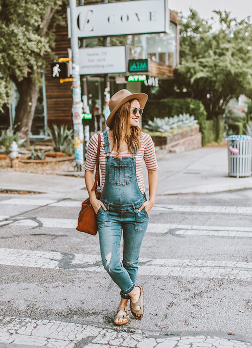 Chica embarazada posando para una sesión de fotos en medio de la calle mientras usa un overol, sandalias y sombrero 