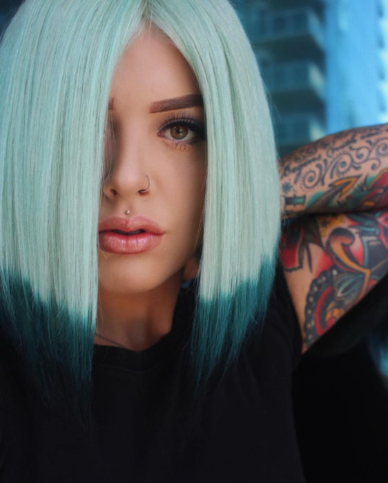 Tinte color verde menta; chica con cabello corto y lacio con tatuajes en los brazos, piercings en la nariz y labio superior 