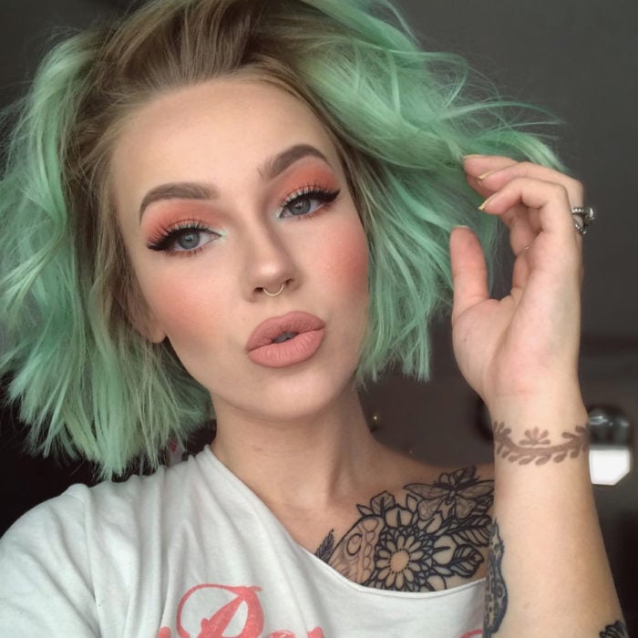 Tinte color verde menta; chica con cabello corto y ondulado con maquillaje natural, tatuaje en la clavícula y piercing en el tabique