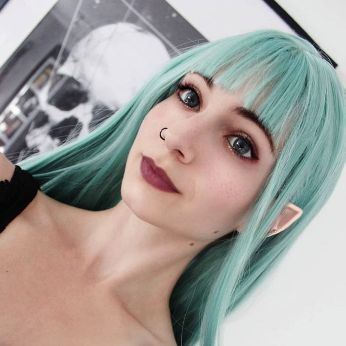 Tinte color verde menta; chica de ojos azules, con orejas de elfo, con piercing en la nariz y cabello largo y lacio con fleco