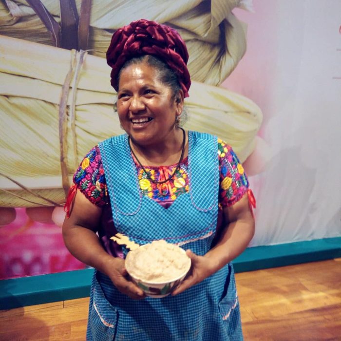 Abigail Mendoza mostrando frente a una pintura de un tamal y con un bowl en la mano