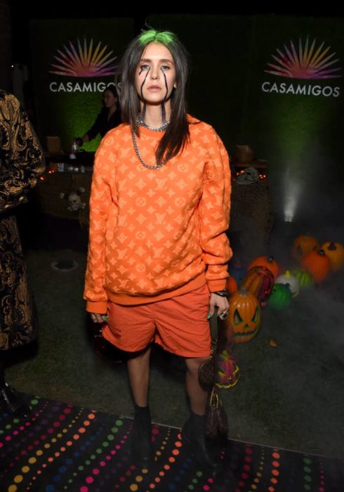 Nina Dobrev disfrazada como la cantante Billie Eilish usando un traje de color naranja 