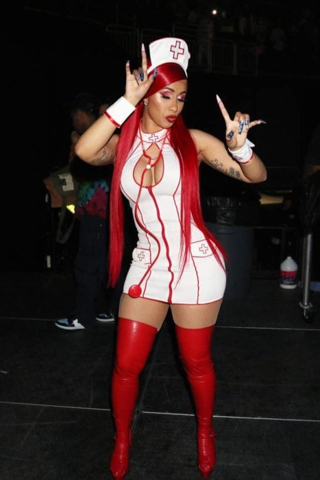 Cardi B disfrazada como una enfermera con un traje blanco, botas y peluca de color rojo 