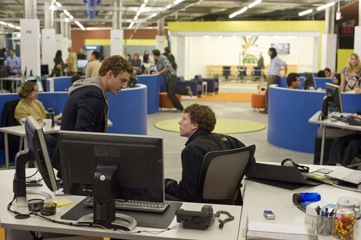 dos hombres hablan, uno sentado frente a una computadora y otro sentado en el escritorio, se ve de fondo una oficina con cubículos azules
