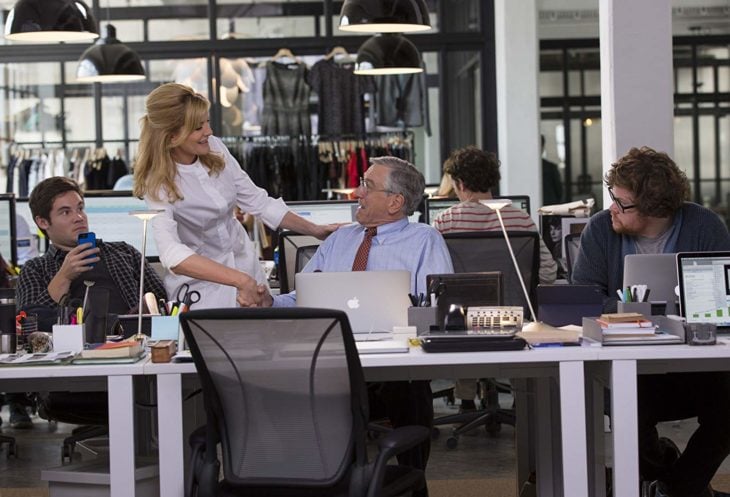 una mujer de pie habla con un hombre que está sentado en un escritorio en donde hay otros dos hombres trabajando