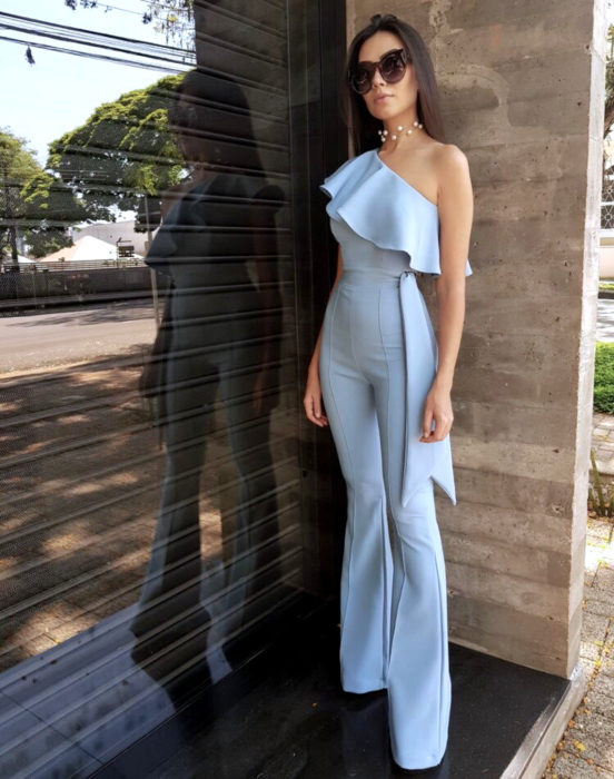 Looks con jumpsuit; mujer de cabello largo, lacio y castaño, con lentes de sol granes, collar de perlas y enterizo elegante color azul cielo de un solo hombro, con holán ancho