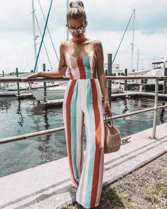 Looks con jumpsuit; chica rubia con peinado de coleta de caballo alta, en un muelle con barcos en el mar; vestida con un enterizo playero de tirantes y rayas anaranjadas, blancas,verdes y rosas verticales, bolsa de mano de yute