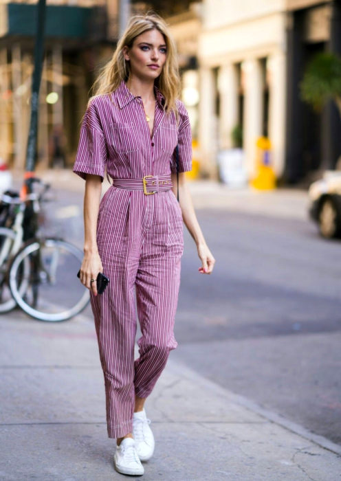 Looks con jumpsuit; chica de cabello rubio, largo y despeinado caminando en la calle con enterizo retro de botones con cinto, mangas cortas, de color morado con rayas blancas horizontales y tenis blancos