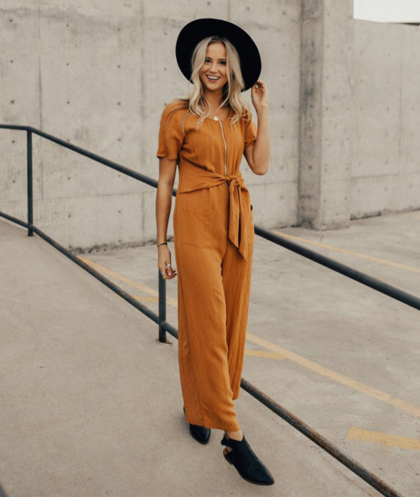 Looks con jumpsuit; chica rubia de cabello largo y ondulado con sombrero negro y enterizo con cierre color anaranjado