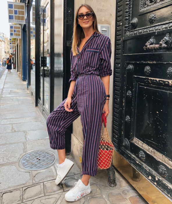 Looks con jumpsuit; chica de cabello castaño, mediado y lacio, posando frente a una gran puerta con ornamentos vestida con un enterizo de mangas largas, botones color morado de rayas blancas verticales y tenis de concha blancos