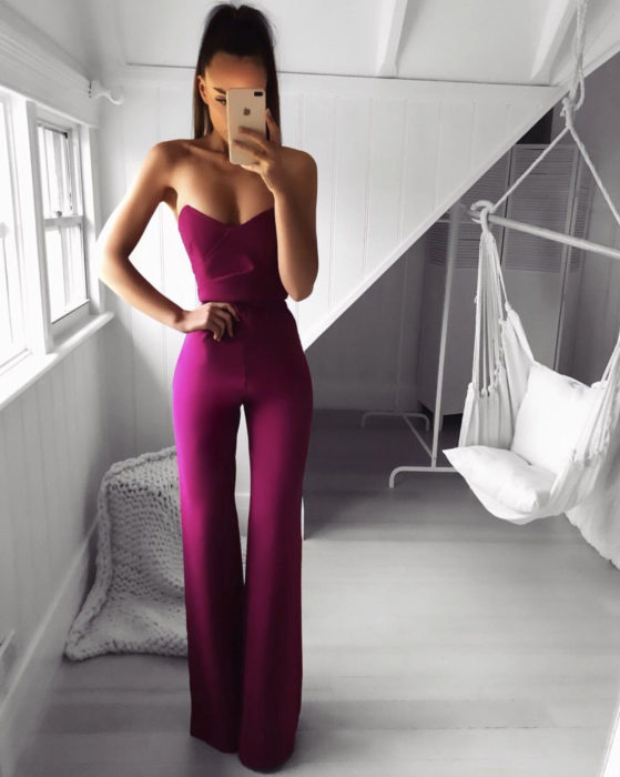 Looks con jumpsuit; chica con peinado de cola de caballo alta, vestida con un enterizo sin hombros color magenta, posando para selfie frente al espejo