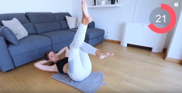 screenshot di un video in cui una donna fa esercizi addominali su una stuoia sul pavimento di legno con un divano grigio dietro