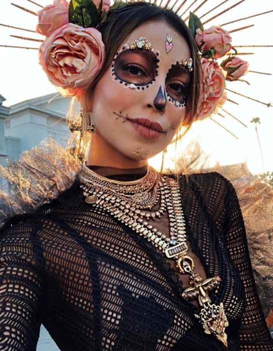 Maquillaje moderno de Catrina; chica disfrazada de Día de Muertos con flores en la cabeza, con collares y aretes de cruz dorada