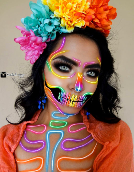 Maquillaje neón moderno de Catrina de Día de Muertos color amarillo, rosa, anaranjado, morado, azul y verde; con flores de cempasúchil en la cabeza; chica de cabello negro a los hombros y ojos verdes claro