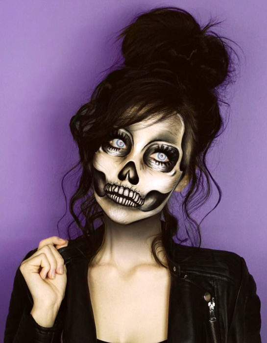 Maquillaje moderno de Catrina realista de Día de Muertos; chica de ojos azules, peinado de chongo alto y despeinado, disfrazada de calavera y chamarra de cuero