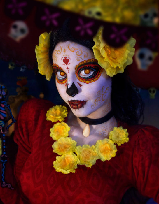 Maquillaje moderno de Catrina de Día de Muertos con flores de cempasúchil amarillas con vestido rojo; mujer disfrazada de El libro de la vida