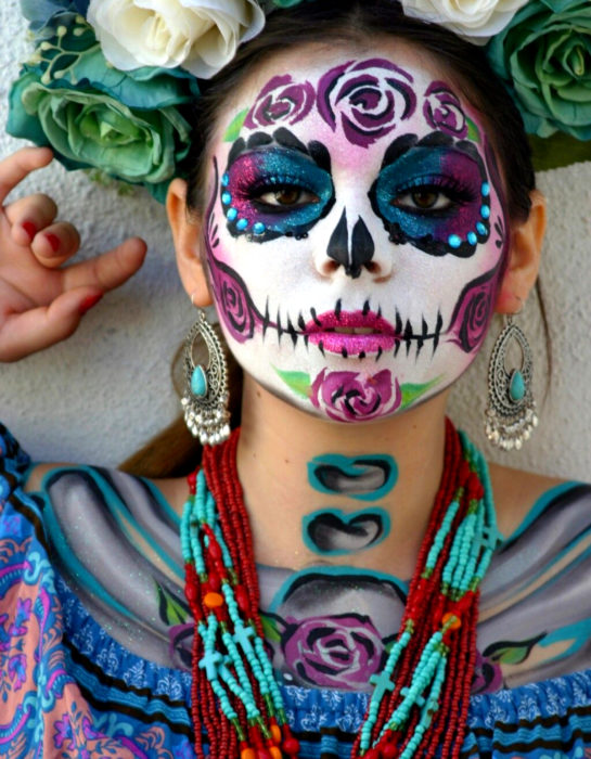 Maquillaje tradicional de Catrina de Día de Muertos de color rosa, azul y morado con glitter, corona de flores blancas y verdes y accesorios típicos