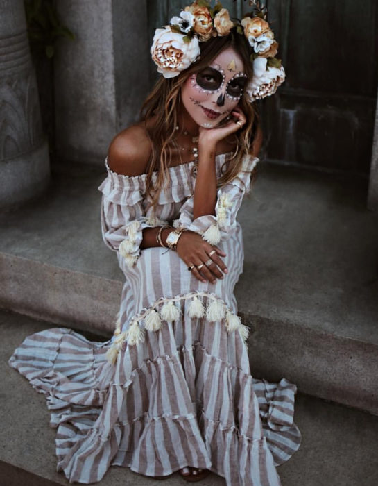 Maquillaje dorado moderno de Catrina de Día de Muertos; chica sentada en escaleras con maxivestido sin hombros de rayas verticales blancas y cafés; flores en la cabeza