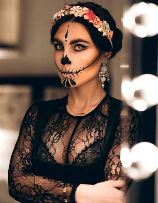 Maquillaje moderno de Catrina de Día de Muertos; mujer de ojos verdes con peinado de chongo bajo elegante, diadema de flores, aretes grandes y blusa de encaje negro