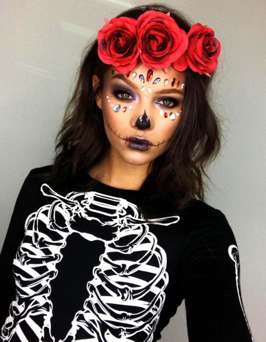 Maquillaje moderno de Catrina de Día de Muertos, sombra de ojos morada y negra, con piedras swarovski en el rostro, con blusa de estampado de calavera, corona de flores rojas