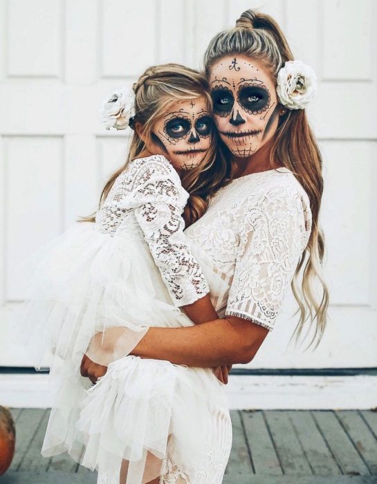 Maquillaje moderno de Catrina de Día de Muertos; madre e hija disfrazadas de calavera con vestidos blancos de encaje y flores en el cabello 