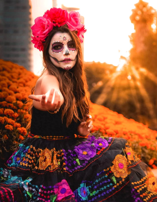 Maquillaje tradicional de Catrina de Día de Muertos con corona de flores en la cabeza y pedrería; chica con falta con flores bordadas en un campo de cempasúchil 