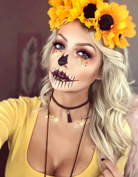 Maquillaje moderno de Catrina de Día de Muertos; chica rubia de ojos azules con corona de girasoles en la cabeza y glitter en el rostro