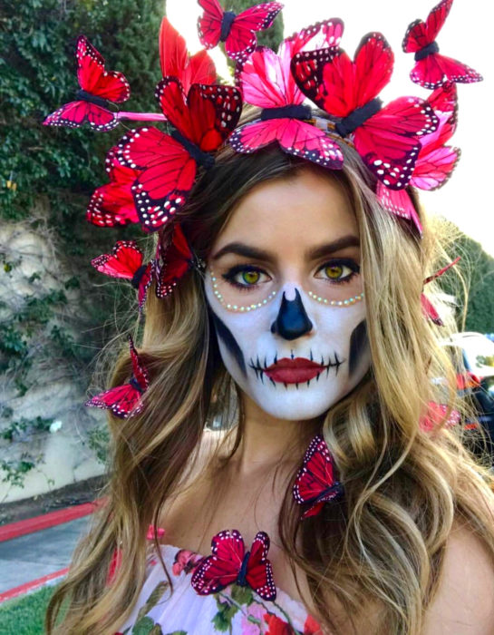 Maquillaje moderno de Catrina de Día de Muertos; chica de cabello largo, rubio y ondulado con corona de mariposas rojas en la cabeza