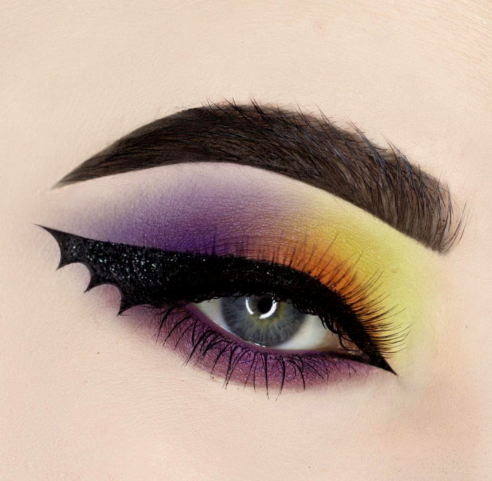 Maquillaje de ojos de Halloween; sombra morada, amarilla y anaranjada con delineado en forma de ala de murciélago 