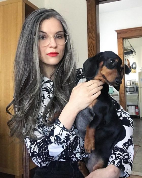 Chica de cabello largo y canoso parada frente a una cámara tomandose una foto mientras está con su perro 