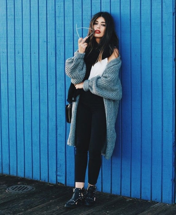 Oversized cardigan; chica de cabello castaño, con suéter holgado tejido color gris con leggins, botines negros con estoperoles, recargada en una pared color azul