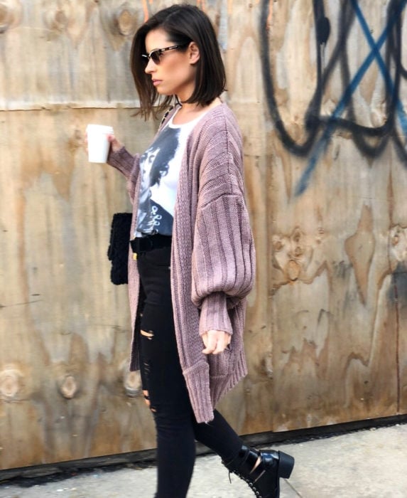 Oversized cardigan; chica caminando en la calle con un vaso de café, mujer de cabello corto, lentes de sol, playera de Prince, suéter holgado rosa tejido