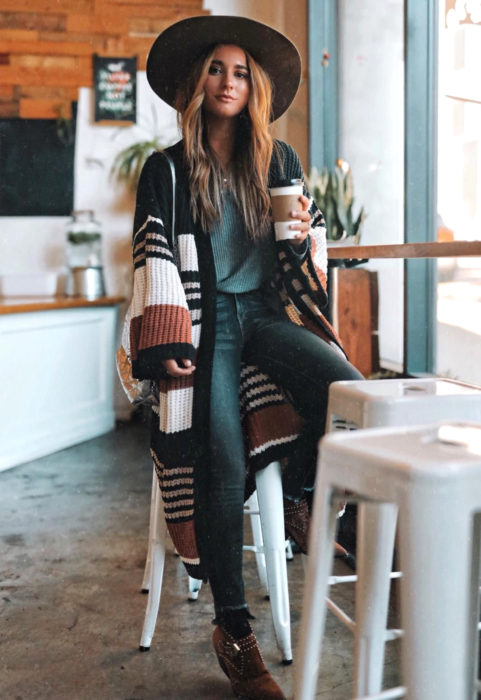 Oversized cardigan; mujer con estilo boho en un café, chica con cabello rubio, sobrero de ala ancha, botines, suéter holgado y largo