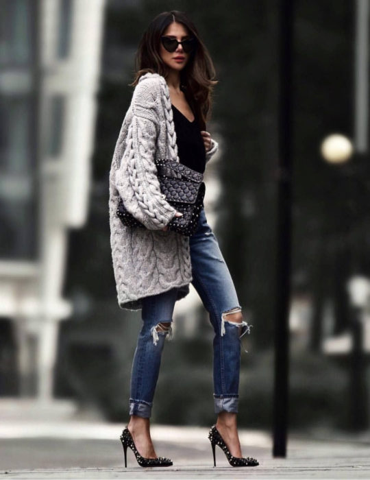 Oversized cardigan; mujer de cabello largo y castaño, con lentes de sol en forma de corazón negros, con pantalones rasgados de la rodilla, zapatillas stiletto y suéter holgado y largo, tejido color gris,bolsa de mano negra con estoperoles 