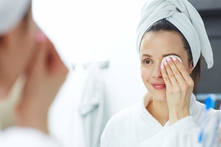 Chica frente al espejo usando una bata de baño y una toalla en la cabeza mientras desmaquilla sus ojos con una almohadilla de algodón 