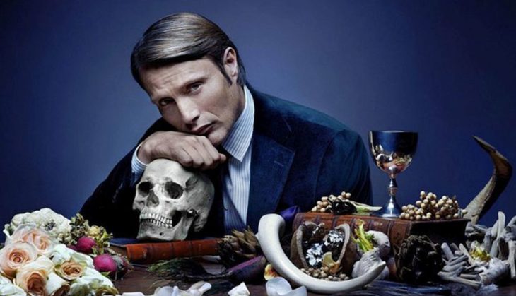 Hombre sentado a la mesa recargado en un cráneo, escena de la serie Hannibal 