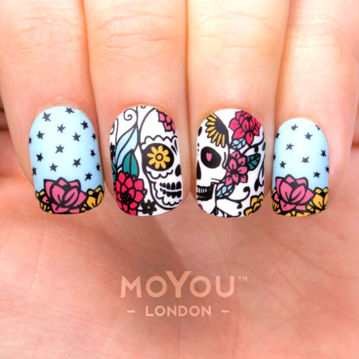 Manicura de Día de Muertos; uñas largas cuadradas pintadas con esmalte azul cielo con diseño de cráneos con flores amarillas, rojas y rosas, con estrellas