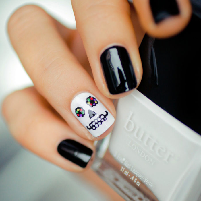 Manicura de Día de Muertos; uñas cortas cuadradas pintadas de negro y una uña con diseño de cráneo con flores en los ojos
