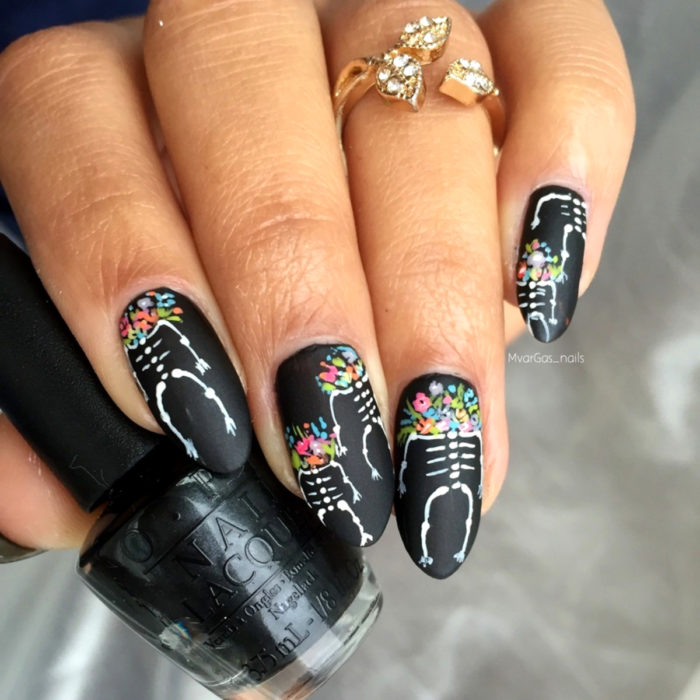 Manicura de Día de Muertos; uñas largas en forma de almendra pintadas de color negro con calaveras y flores de colores rosas, azules, amarillas, moradas y moradas